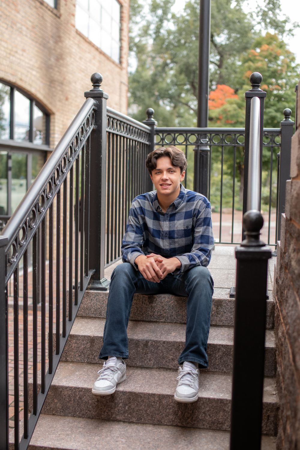A person in a plaid shirt sits casually on concrete steps with black metal railings near a brick building.