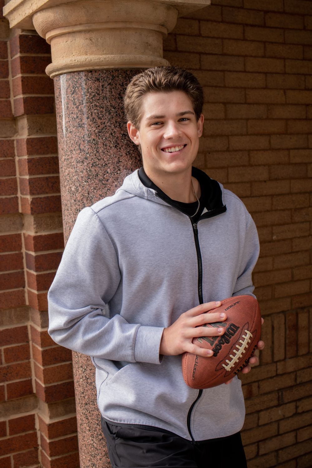 A person in a gray zip-up hoodie poses with a football against a brick wall background.