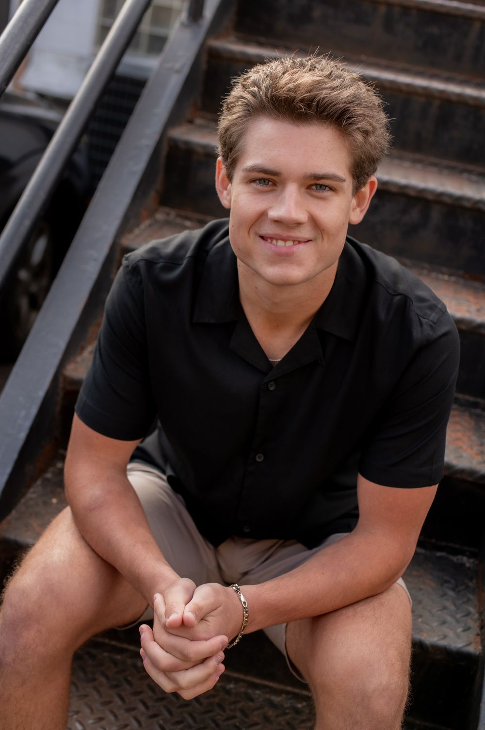 Young person in a black polo shirt sitting on metal stairs outdoors with a warm smile.
