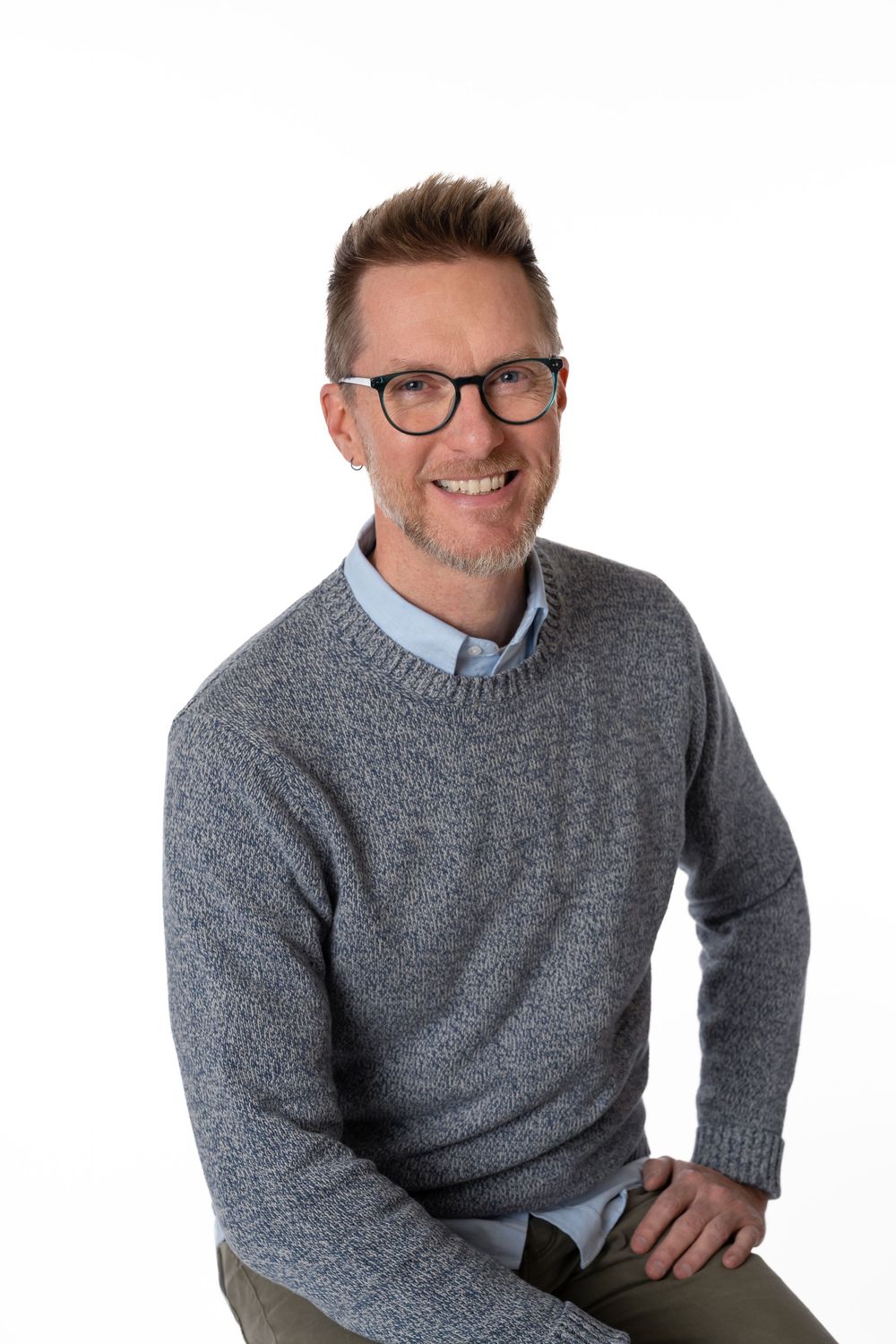 Smiling middle-aged man with glasses wearing a gray sweater over a blue shirt, seated against a white background.