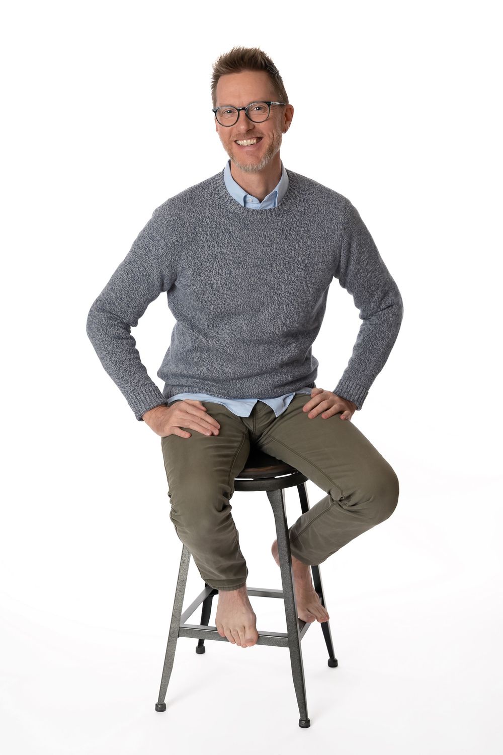 Smiling man in glasses, gray sweater, and olive pants sits barefoot on a metal stool against white background.