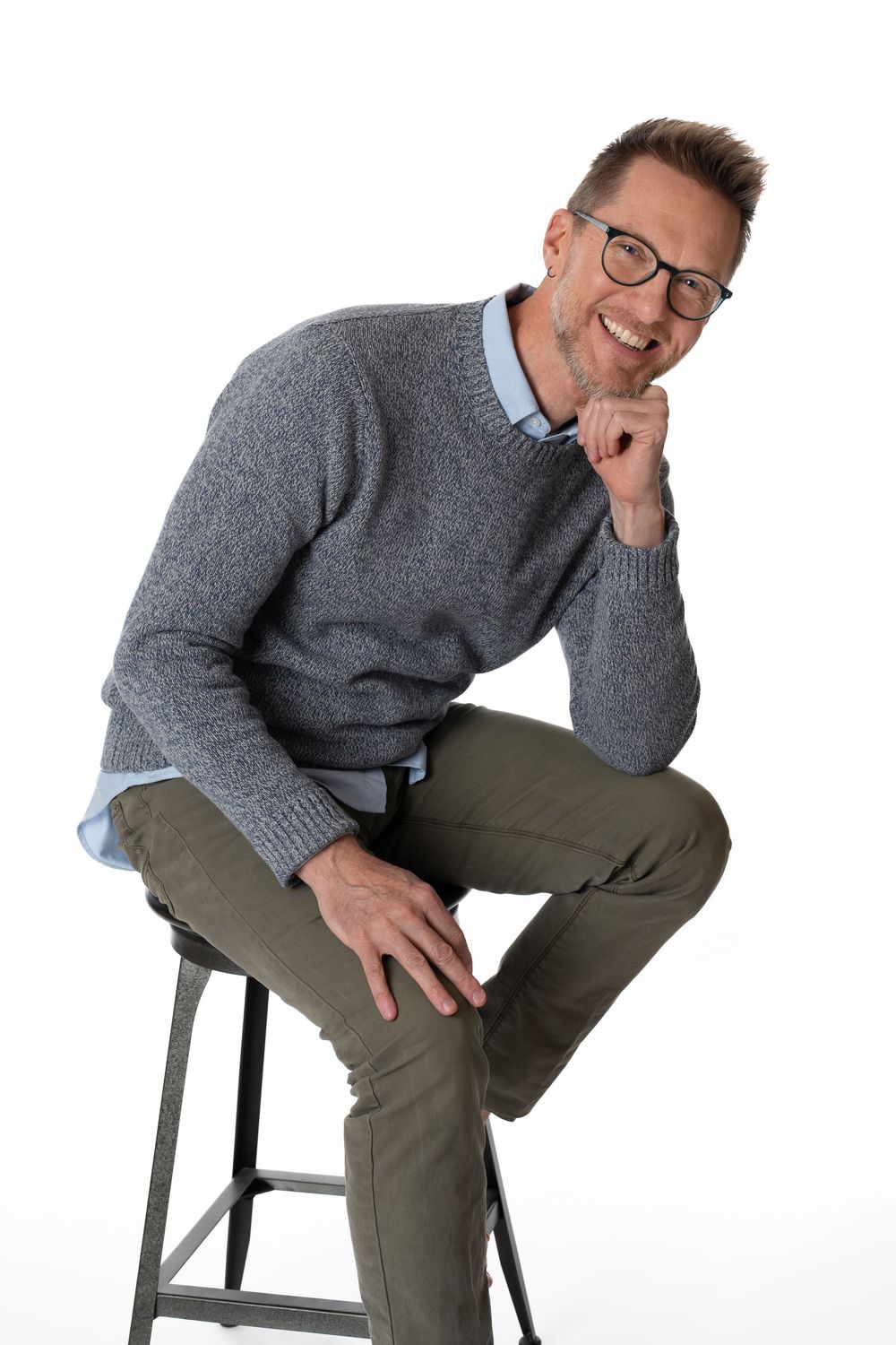 Man in gray sweater and olive pants sits on stool with hand on chin, smiling against white background.