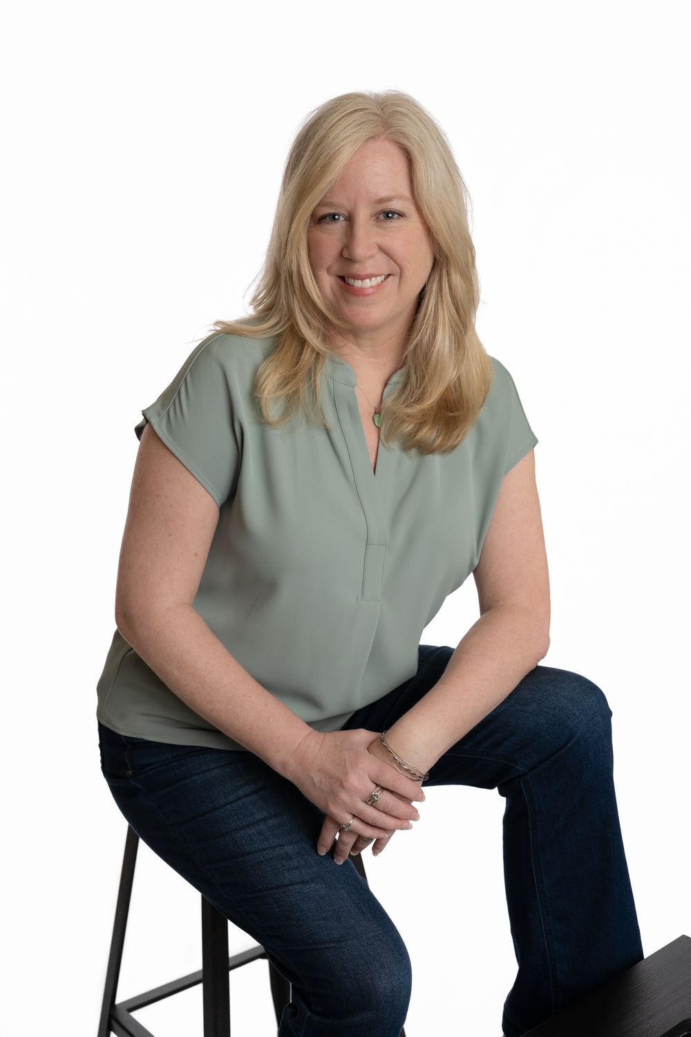 Blonde woman in sage green top and jeans sitting on stool smiling against white background.