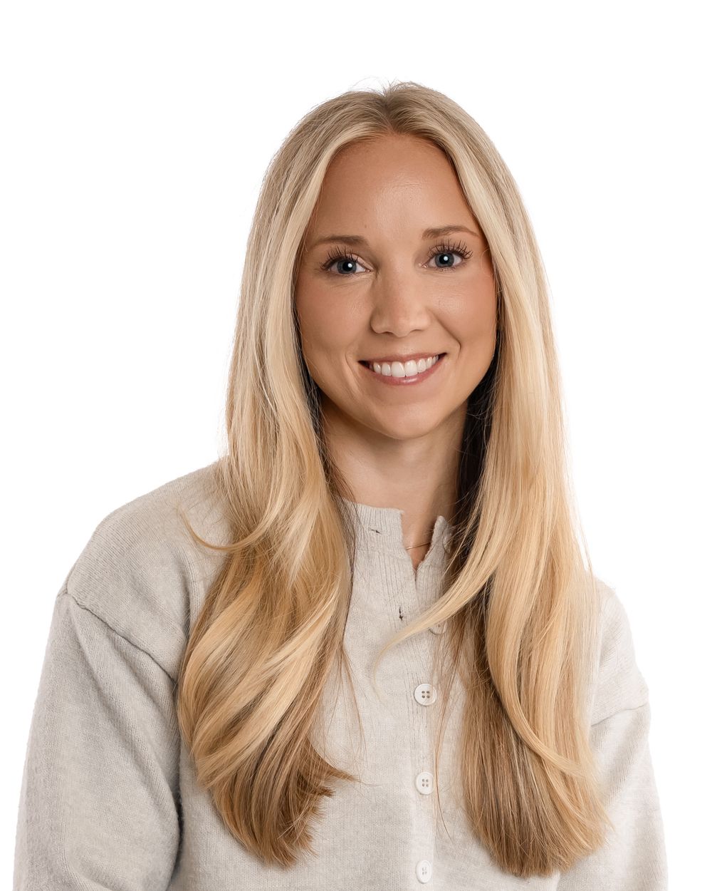 Smiling blonde woman sitting on a wooden stool, wearing a beige sweater, black pants, and brown flats against white background.