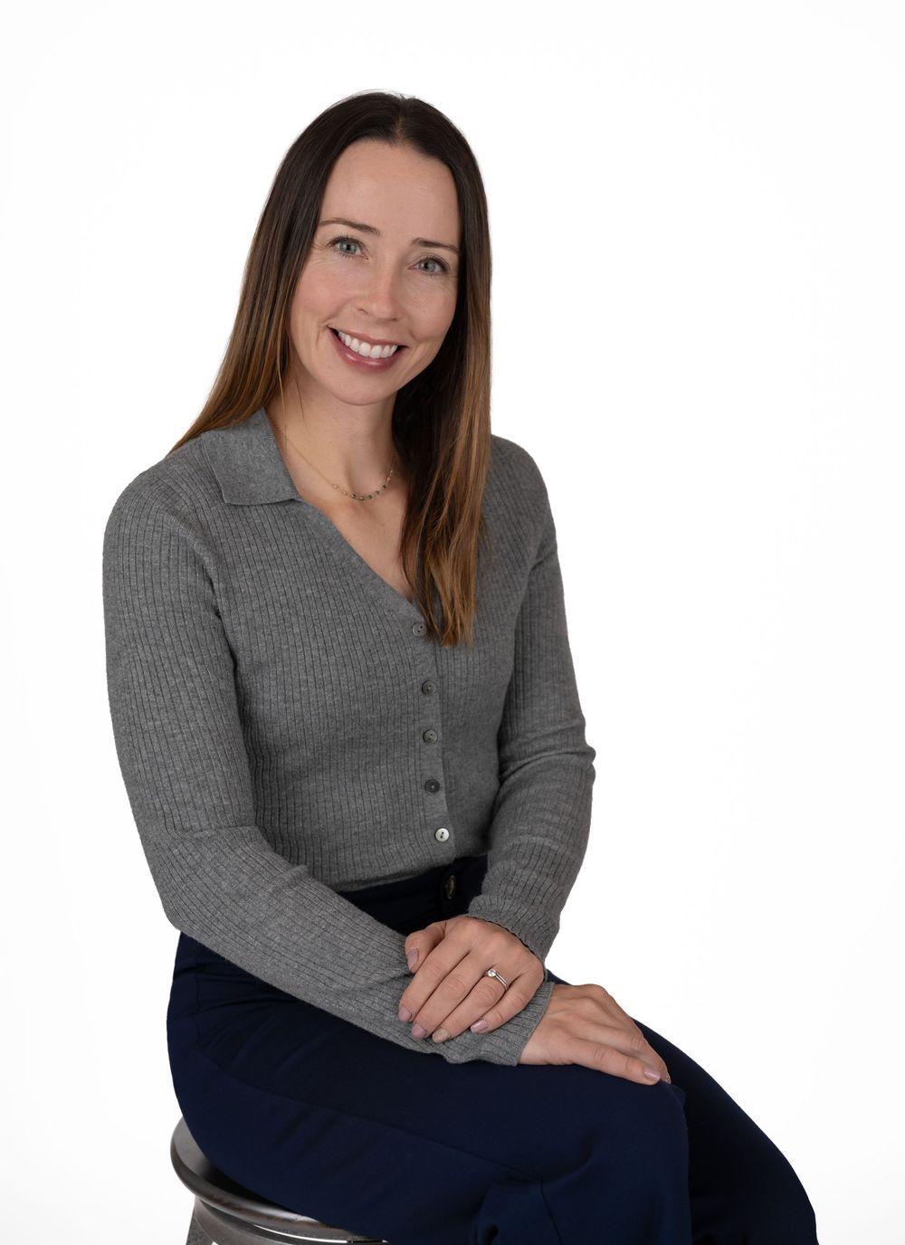 Professional woman in gray cardigan sitting on stool, smiling, with long brown hair against white background.