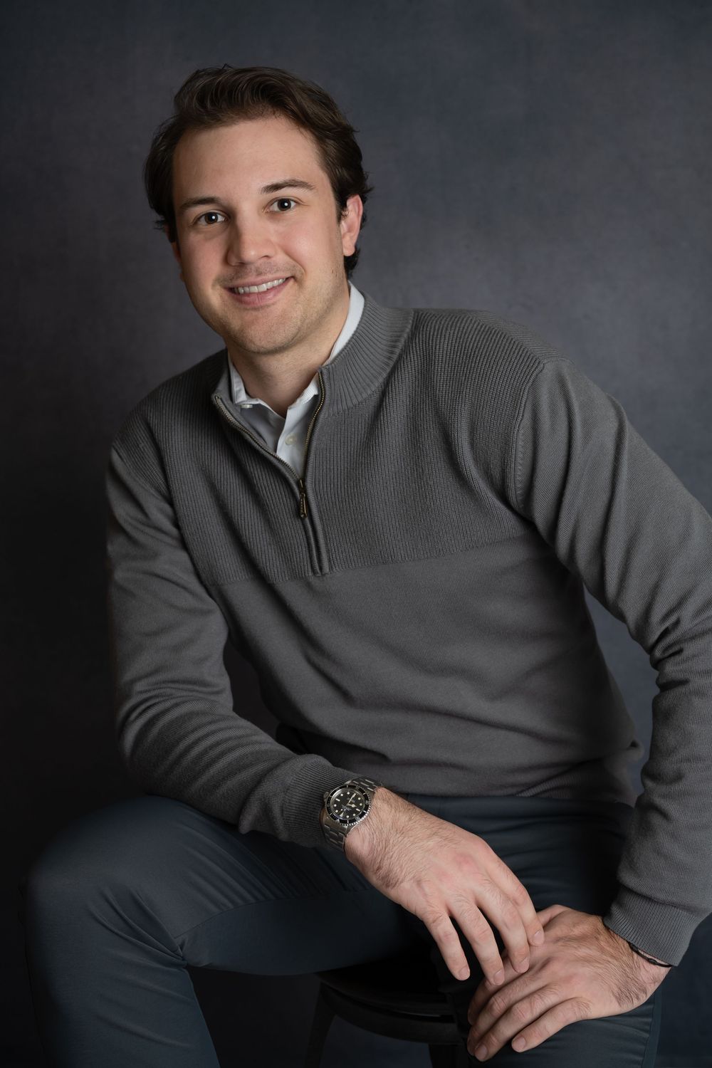Smiling man in gray quarter-zip sweater seated casually on stool against dark studio background.
