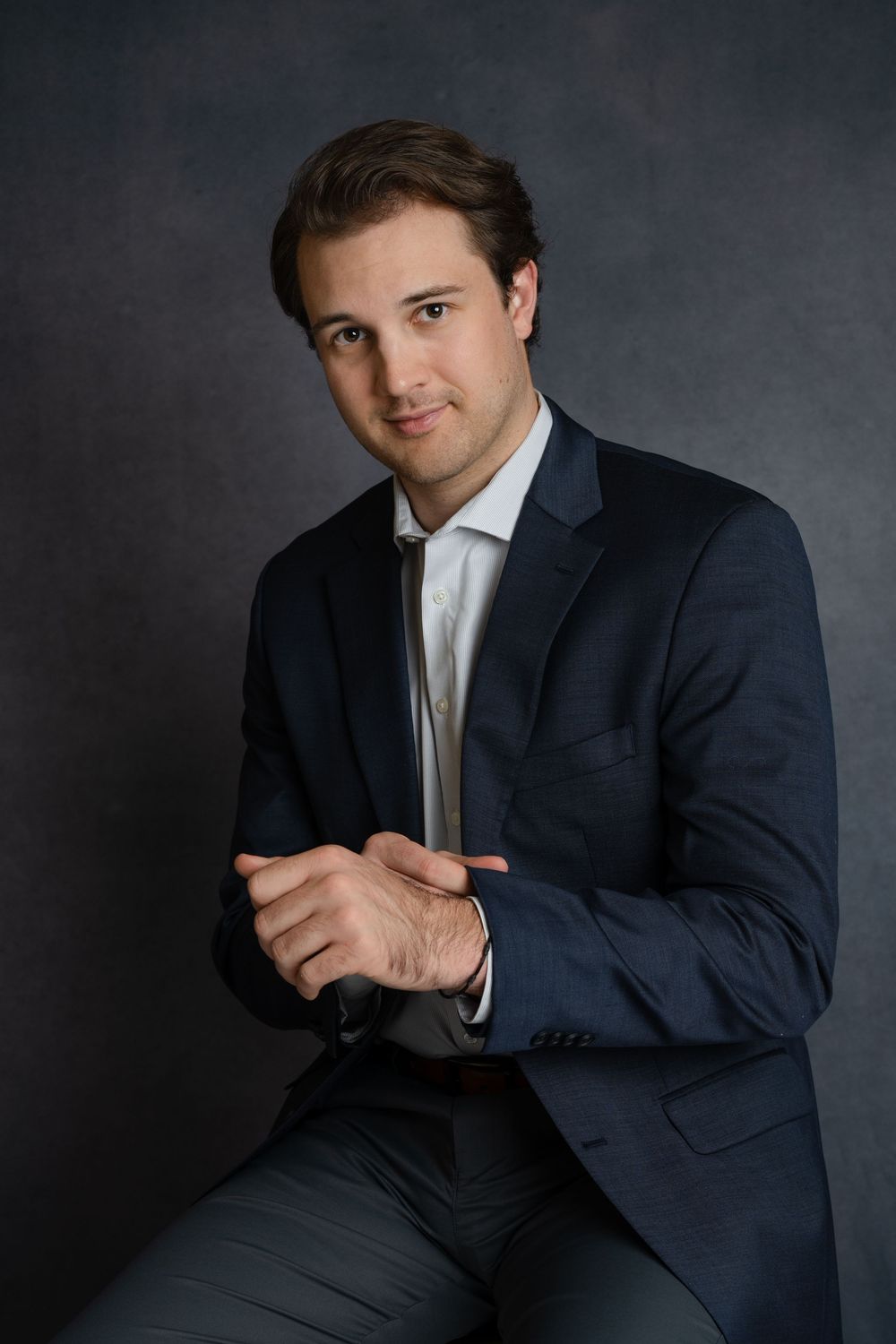 Confident young man in navy suit seated with hands clasped against dark gray studio backdrop.