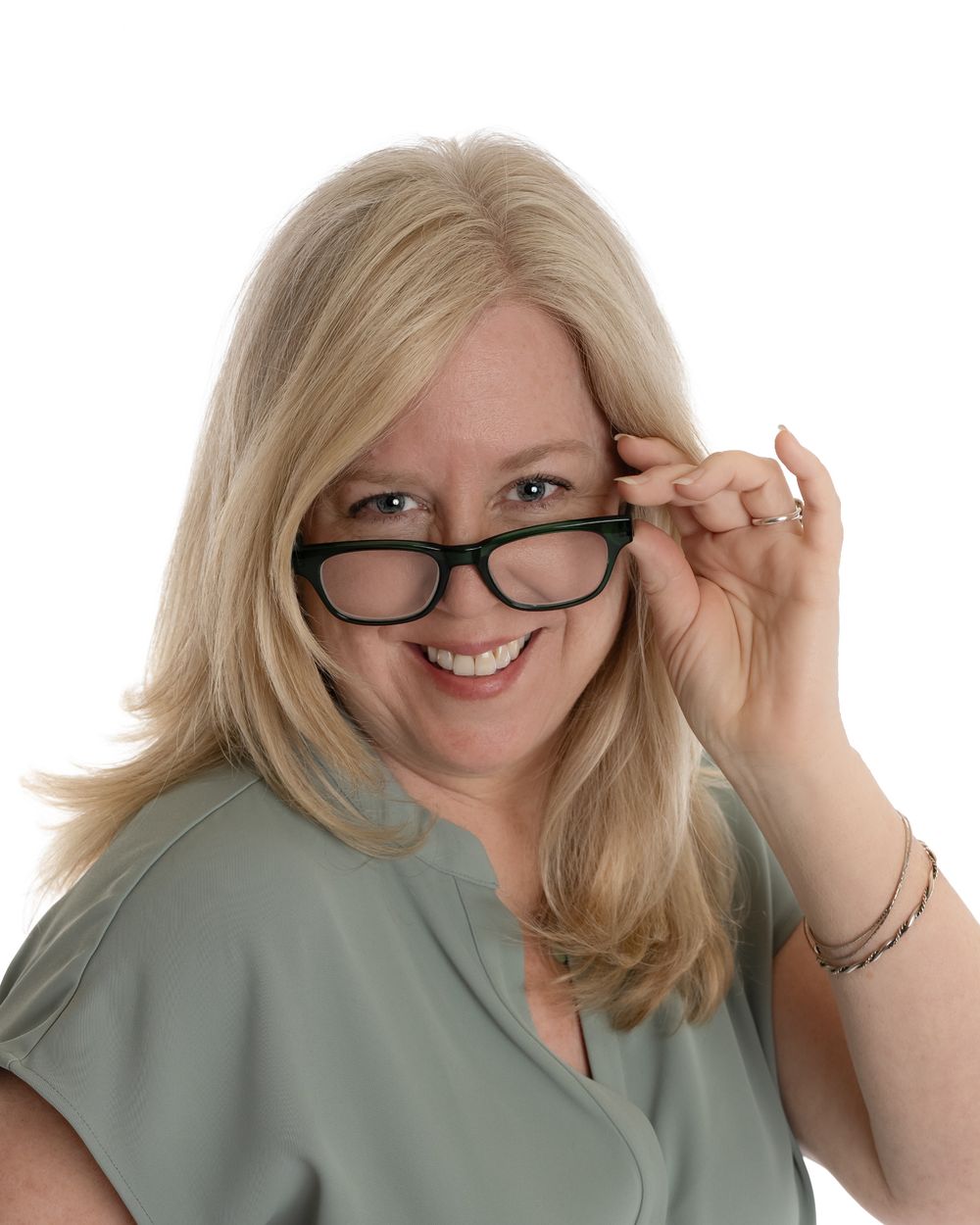 Close-up of smiling blonde woman in sage blouse playfully lowering black-rimmed glasses, white background.