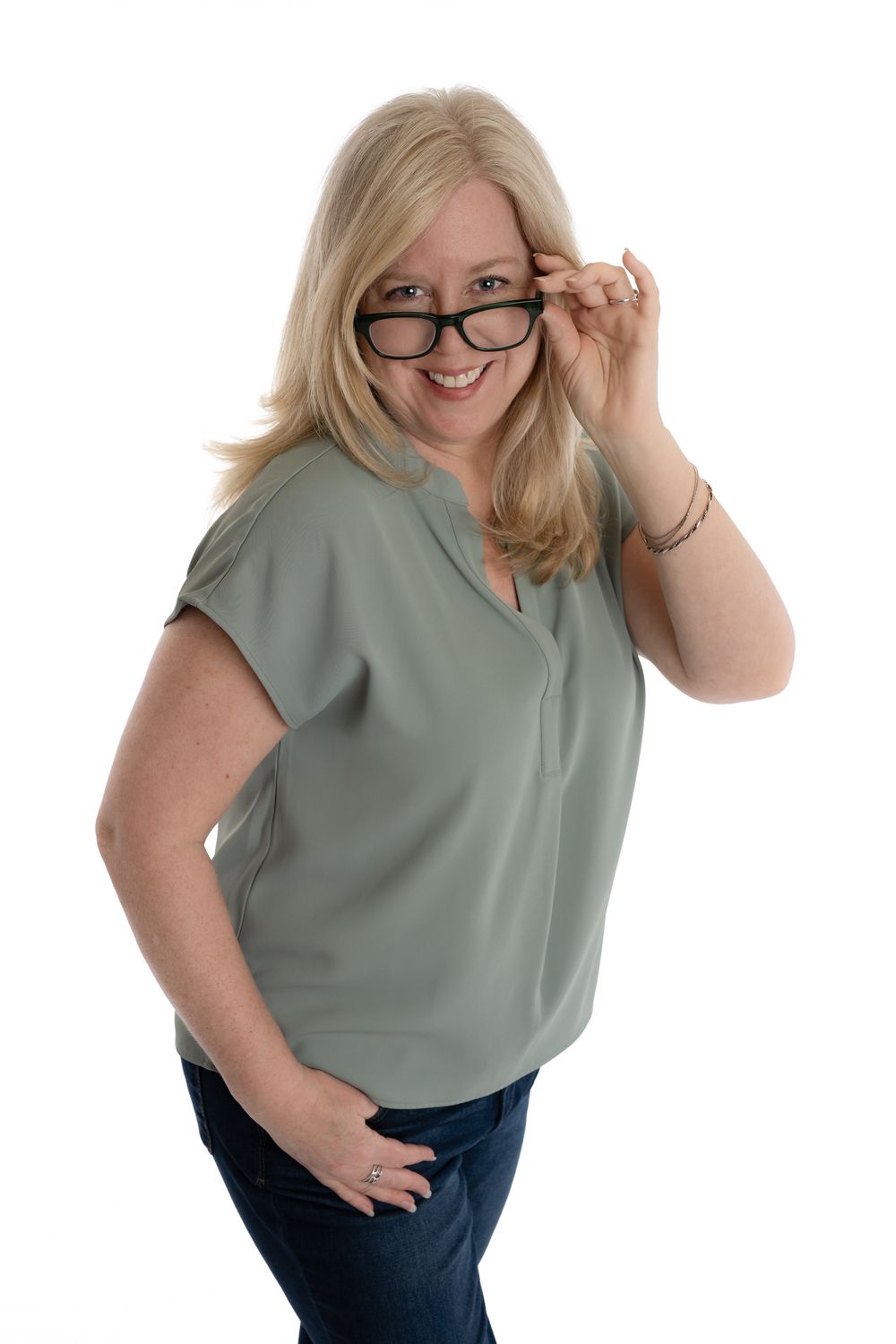 Smiling blonde woman in sage green top pulling down black glasses, posing against white background.