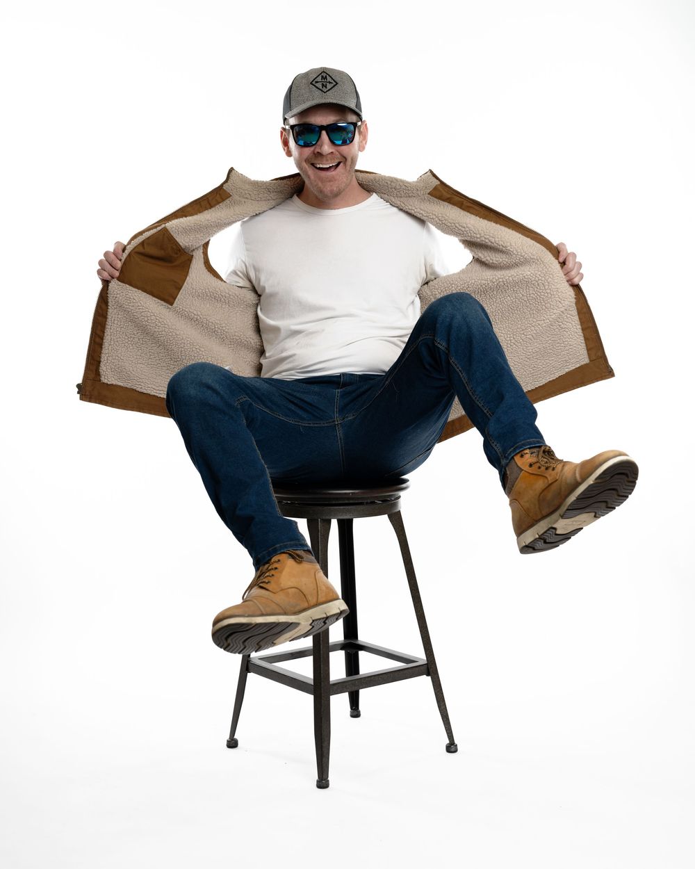 Happy man in white tee and jeans opening sherpa-lined brown jacket while seated on stool.