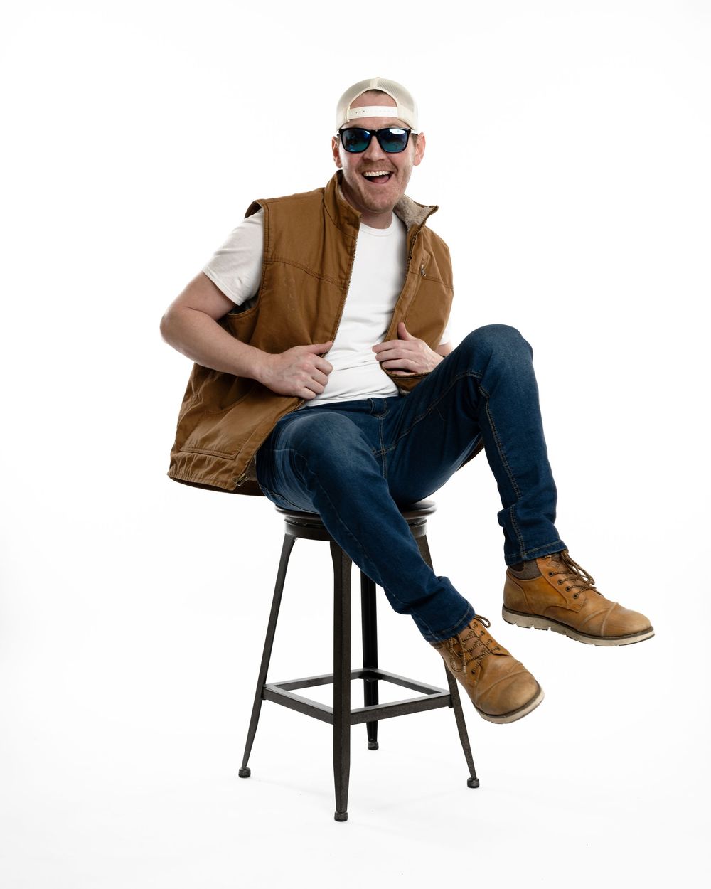 Laughing man in brown vest, white tee, jeans, and sunglasses sitting on metal stool.