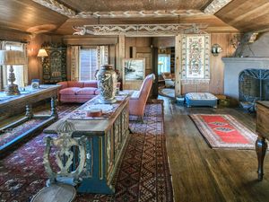 Rustic alpine chalet interior with ornate painted furniture, Persian rugs, wooden beams, and a traditional tiled fireplace.