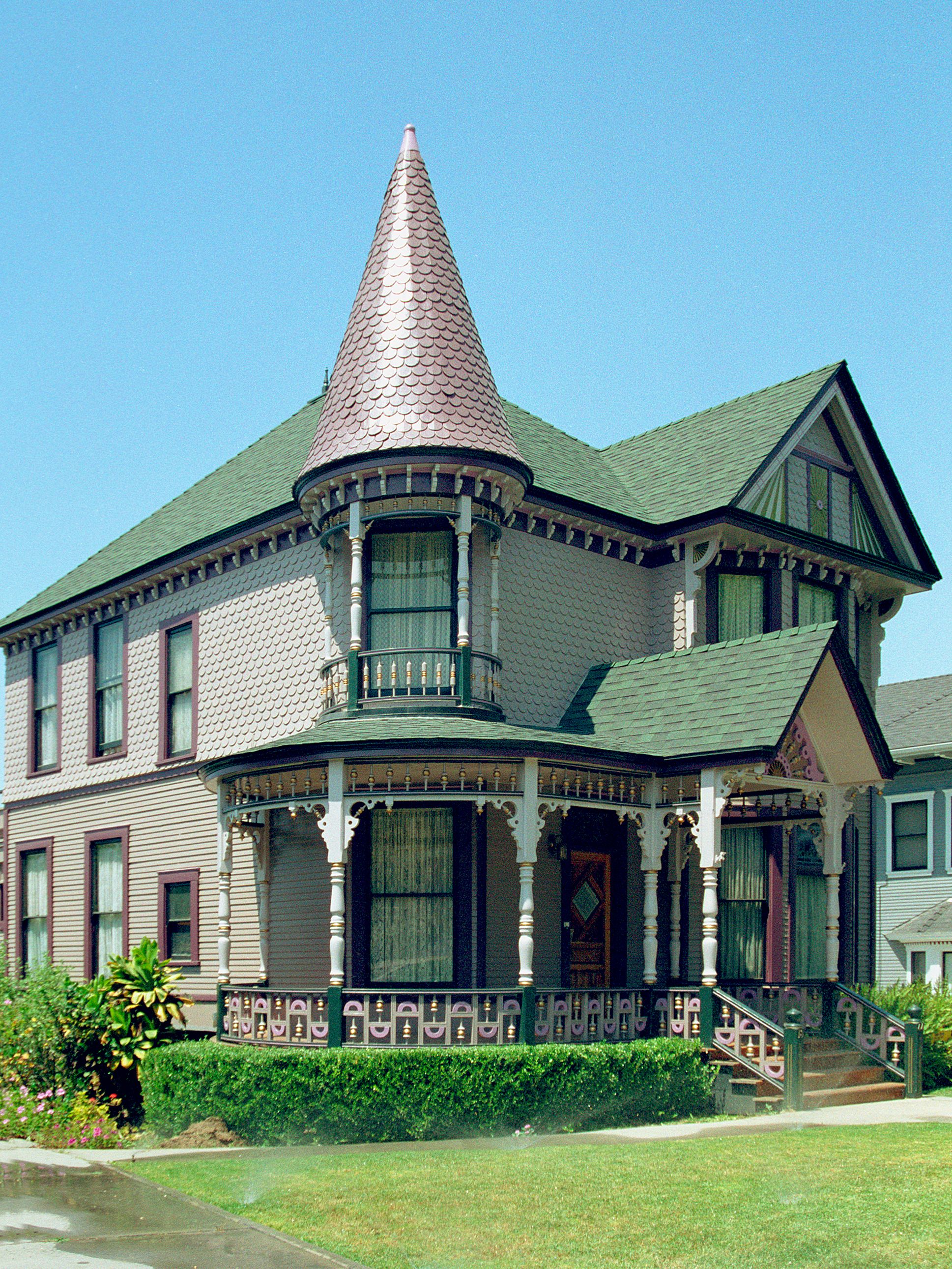 Victorian Queen Anne house with copper spire tower, wrap-around porch, ornate trim, and green roof in a sunny residential setting.