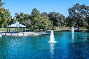 Two fountains spray water in a calm blue park pond with a white gazebo on a wooden dock and tall shade trees along the shore.