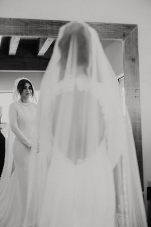 A bride in a long sleeve wedding dress stands near another figure wearing a white veil in a black and white photo.