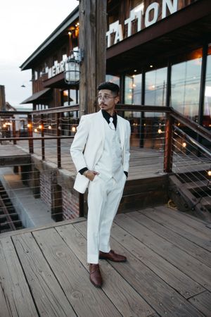 Groom in crisp white suit poses on wooden deck outside Merryman Station.