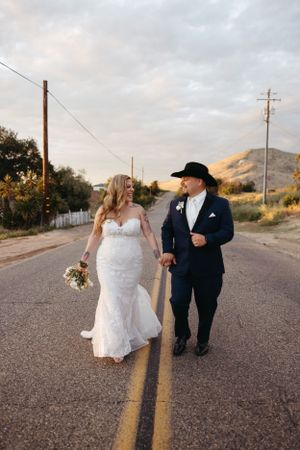 Newlyweds walk down the middle of a rural road at sunset with mountains in background.