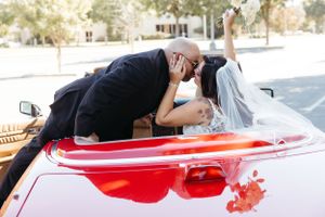 A romantic kiss over a classic red convertible car in a parking lot.