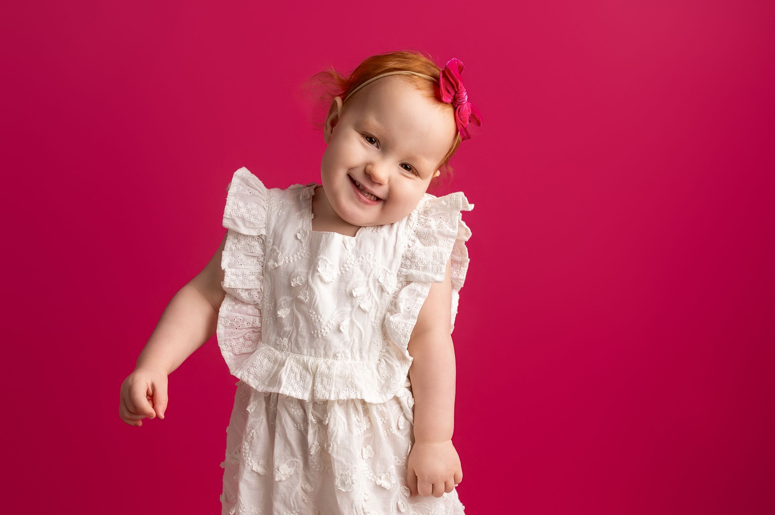 A baby in a white lace dress smiles and poses against a bright pink background.