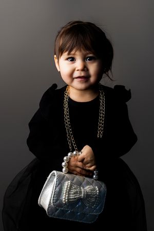 Adorable toddler in black dress poses with silver handbag in dramatic studio lighting sequence.