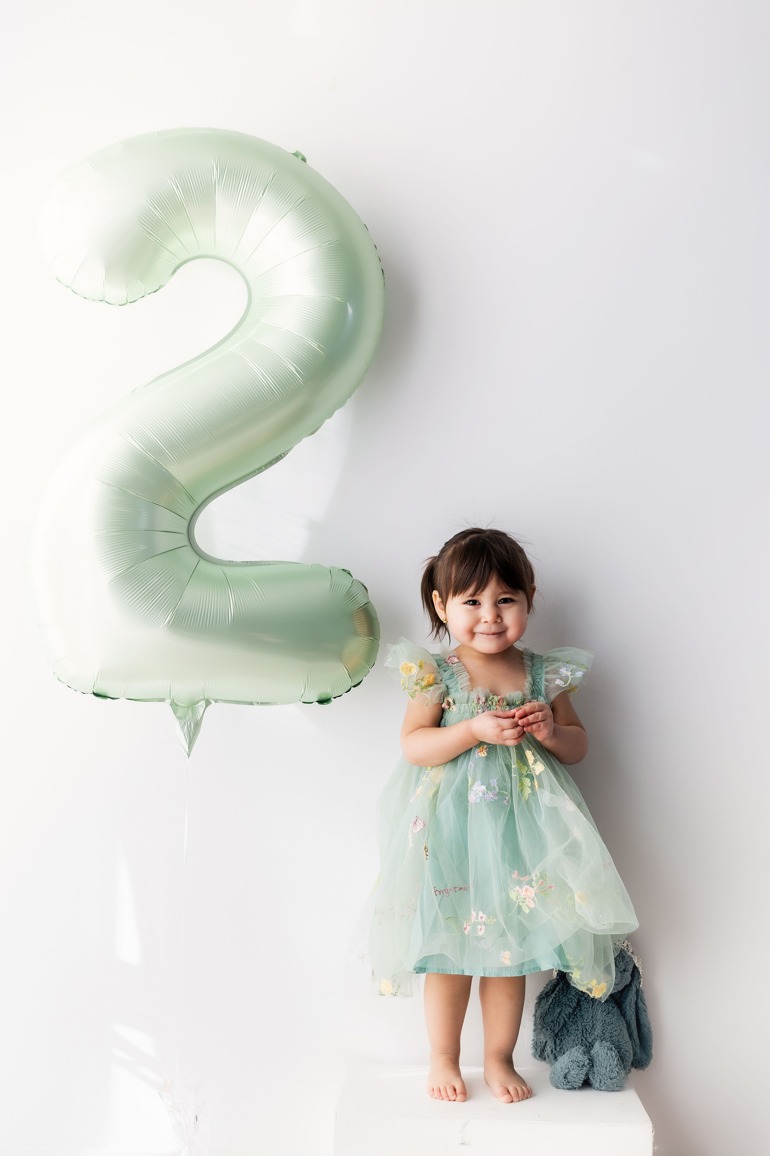 A child in a mint green dress stands next to a large number 2 balloon against a white wall.