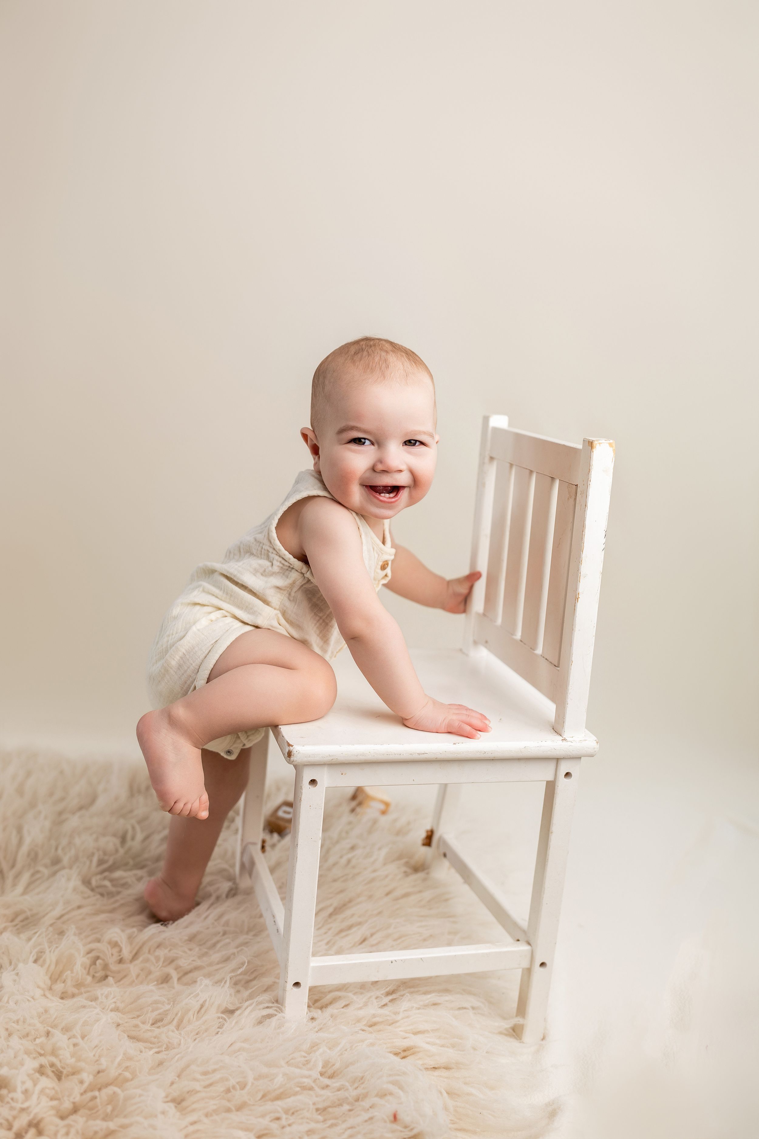 Baby in white diaper plays on white wooden chair against neutral background with fluffy carpet.