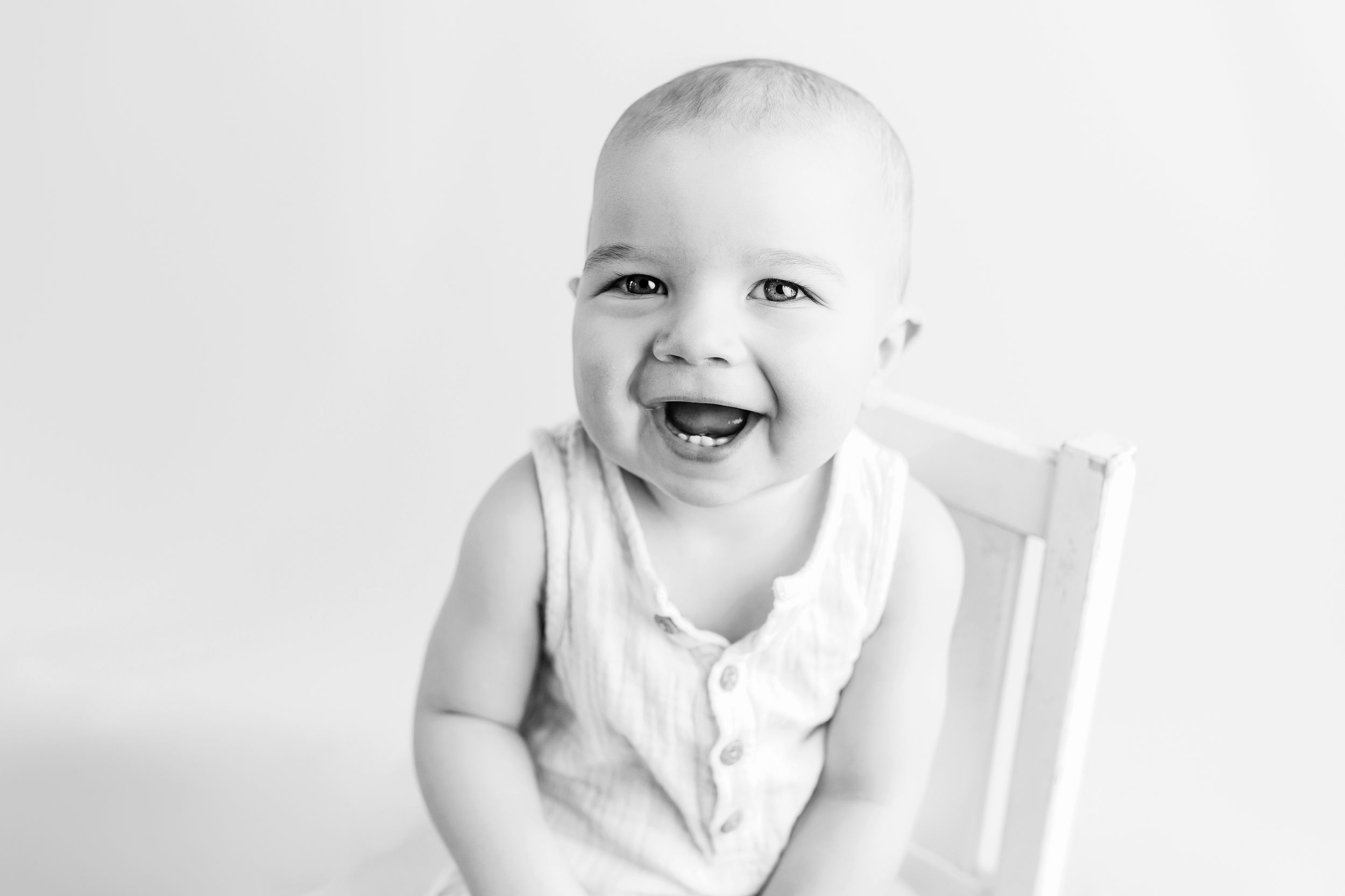 A laughing baby wearing a white onesie sits in a white wooden chair against a light background.