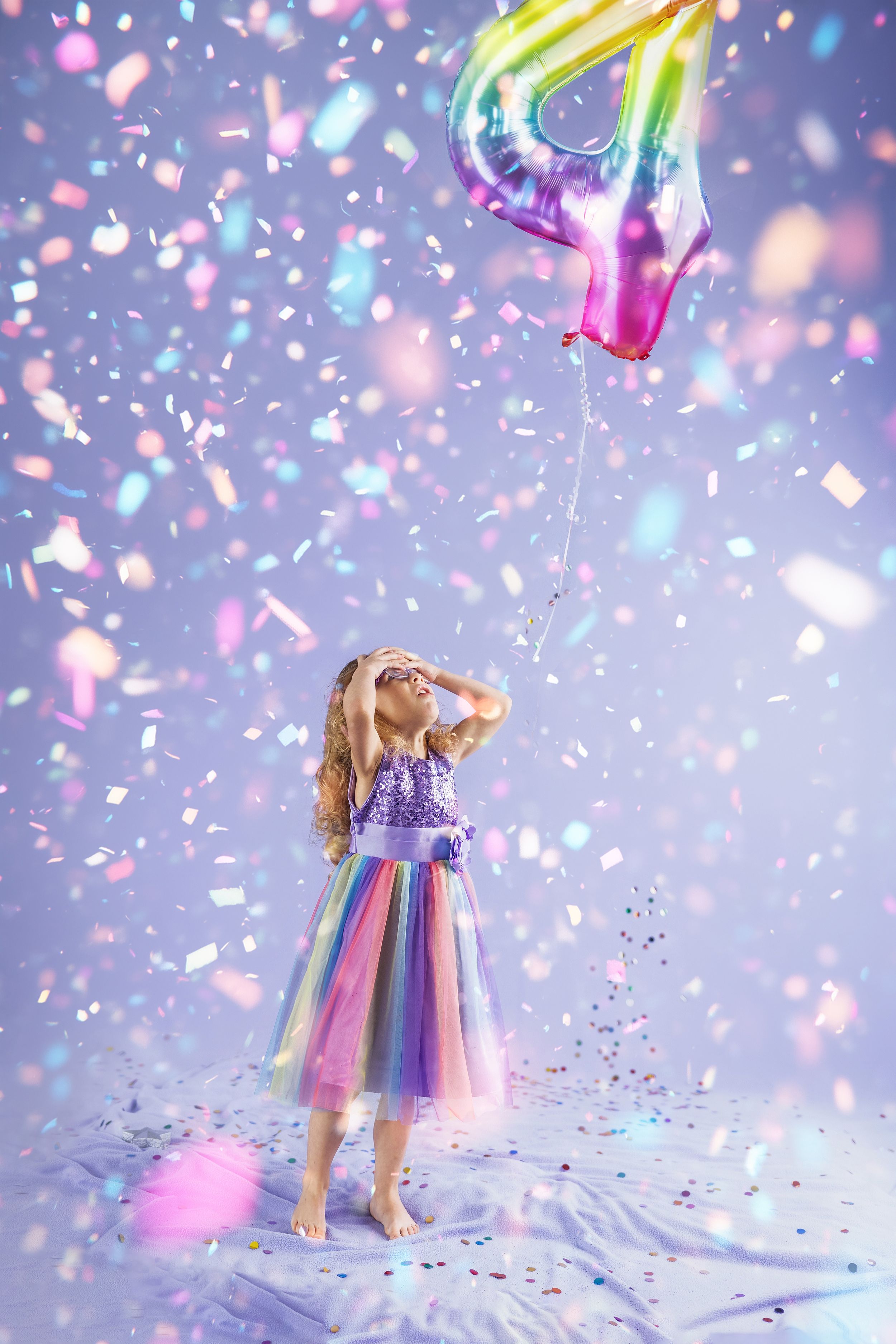 A child in a rainbow tulle dress stands under falling confetti and a colorful balloon in a dreamy purple setting.