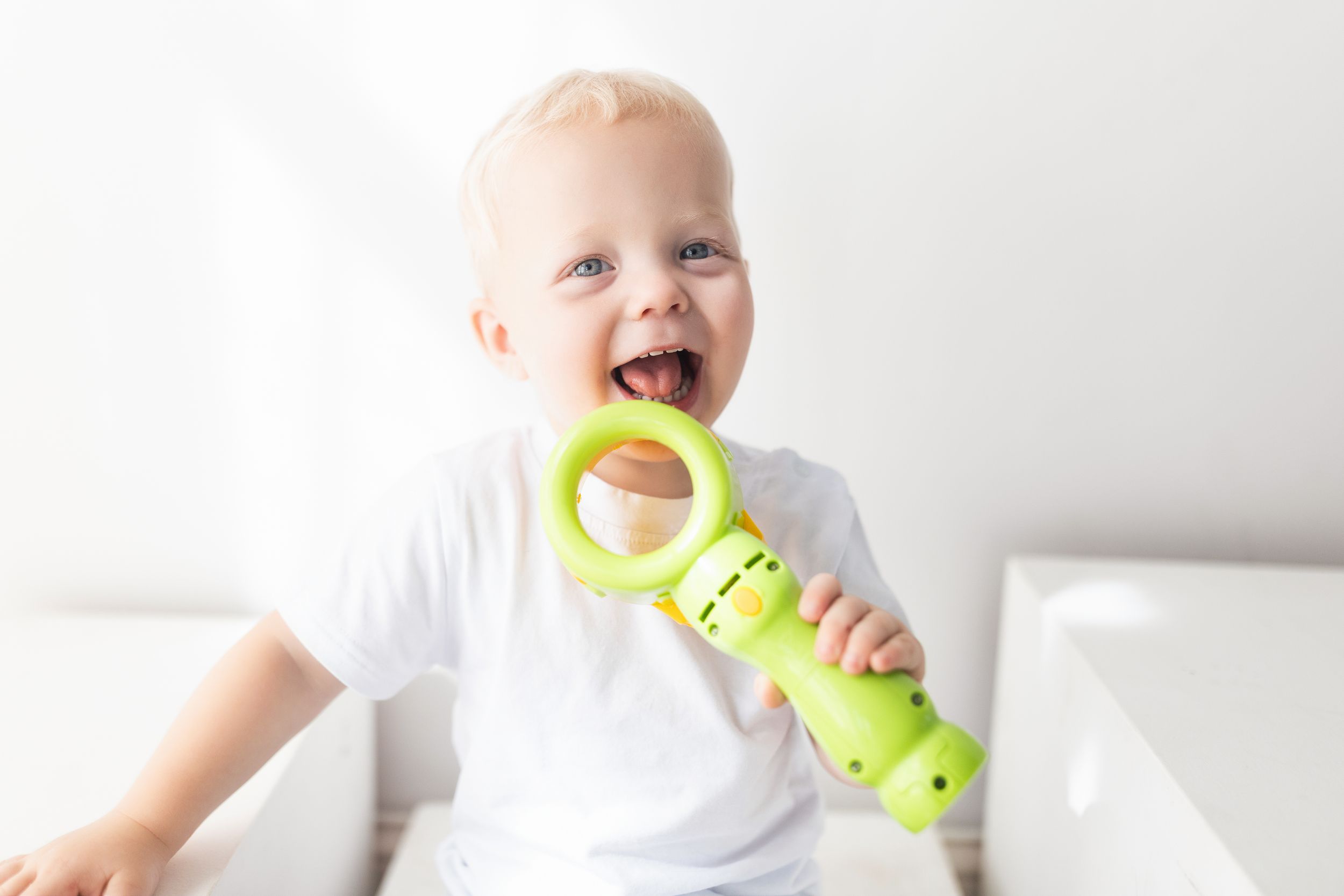 Baby in white clothing holds and chews on a bright yellow teething toy while sitting on white bedding.