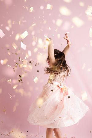 A figure in a flowing white dress twirls joyfully as gold and silver confetti sparkles and falls around them in a pink room.