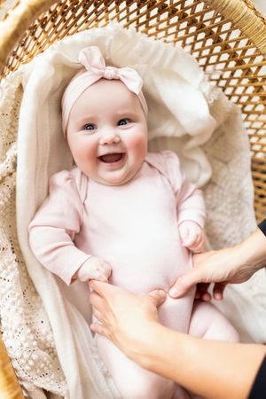 A smiling baby wearing a pink outfit and bow lies in a woven basket lined with soft white fabric.