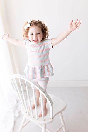 Energetic toddler in pink and blue striped dress playing on white chair.