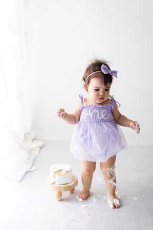 Baby in purple tutu dress taking first steps on white background.