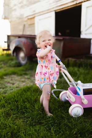 Small feet in a pink patterned dress walking on grass with a white and purple toy stroller.
