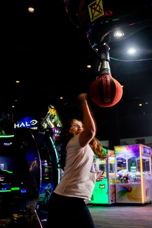 Person in white t-shirt reaches up to hit a punching bag game in an arcade with neon lights and gaming machines.