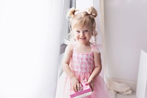 Toddler wearing pink dress posing near window in bright white room.