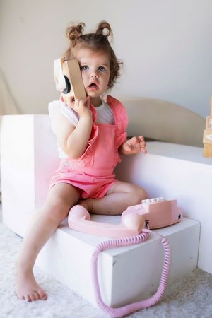 A child plays with a pink vintage toy telephone while sitting in white decor wearing pink romper.