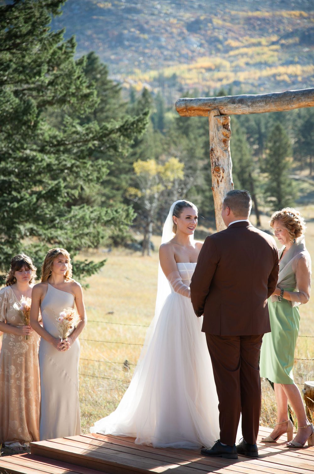 Outdoor mountain wedding ceremony with rustic wooden arch and attendants in pastel dresses.