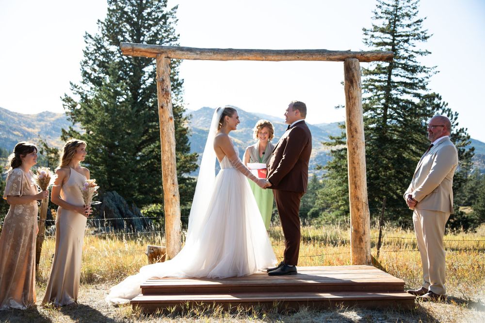 Wedding ceremony under wooden arch with mountain views and pine trees in background.