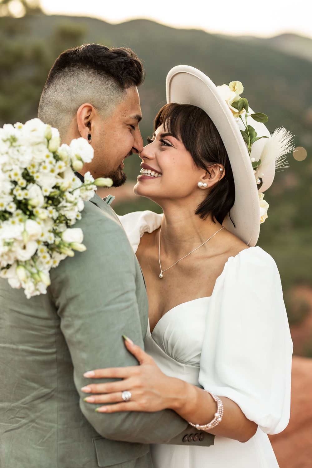 Joyful wedding portrait with bride in white hat and groom holding floral bouquet against nature backdrop.