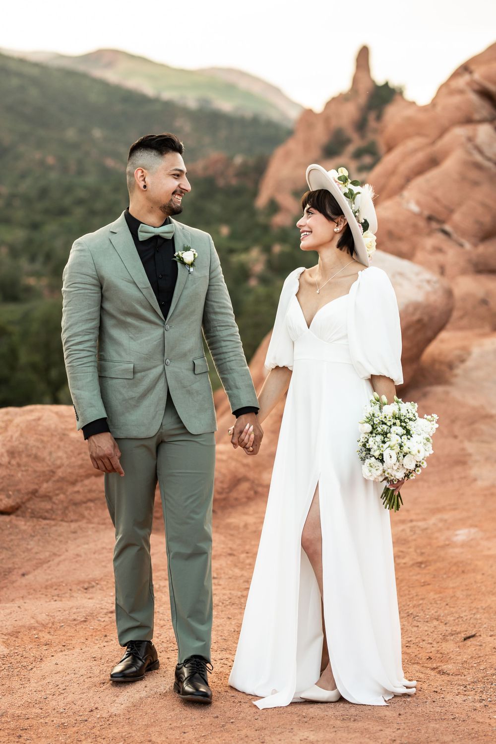 Elegant couple in gray suit and white dress holding hands against red rock landscape.