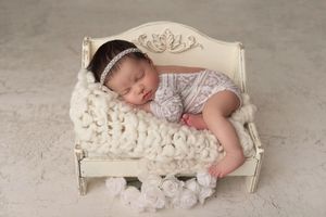 Newborn girl in a white lace outfit sleeping on a tiny white daybed.