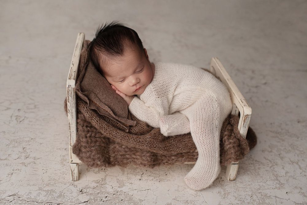 Sleeping newborn boy in a white knit sleeper posed on a white tiny bed with a brown blanket and pillow.