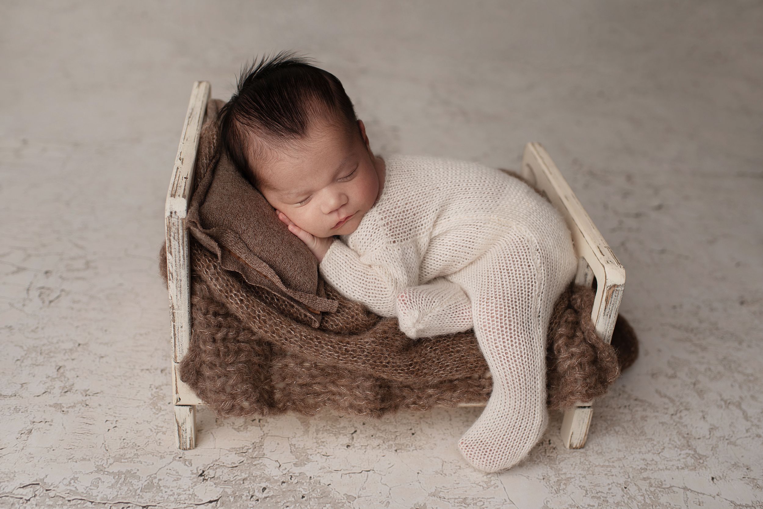 Sleeping newborn boy in a white knit sleeper posed on a white tiny bed with a brown blanket and pillow.