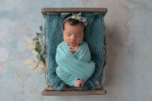 Newborn girl with dark hair in a teal swaddle sleeping on a gray tiny bed on a teal and gray floor drop.