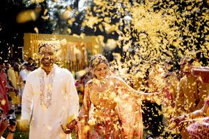 A couple in traditional Indian wedding attire celebrates amidst a shower of golden confetti in an outdoor setting.