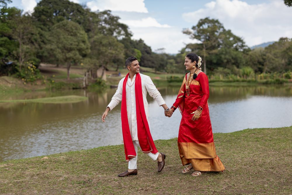 Two people in traditional red and gold wedding attire walk hand in hand by a serene lake surrounded by green landscape.