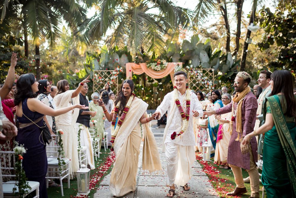 Celebratory wedding moment at an outdoor tropical venue with palm trees and guests in traditional Indian attire.
