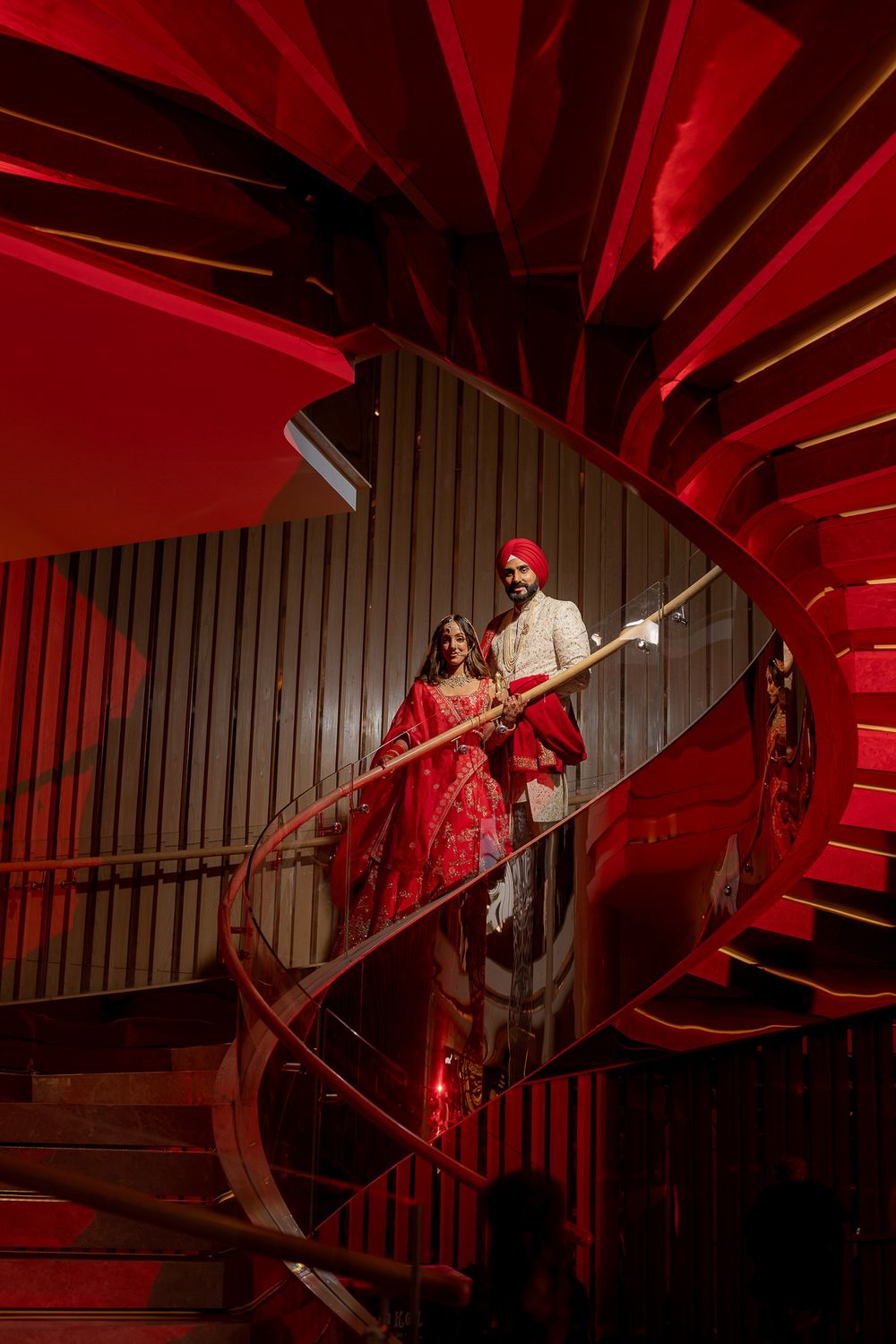 A couple poses on a dramatic spiral staircase illuminated by striking red lighting in a stunning architectural setting.