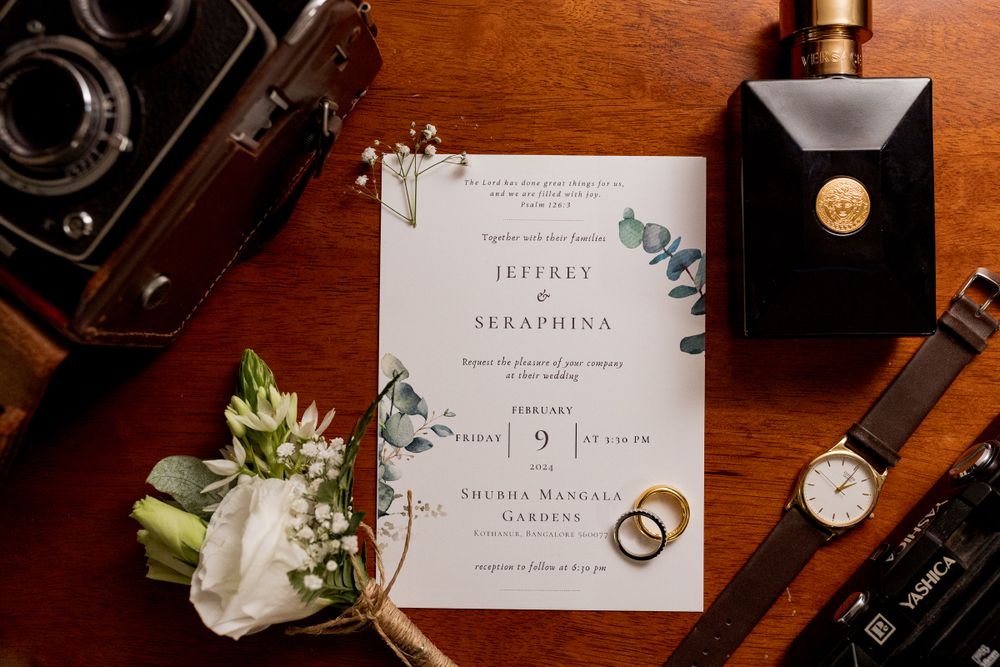 Wedding invitation with floral design displayed with rings, watch, white rose bouquet and vintage camera on wooden surface.